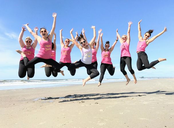 Eight women in matching pink tank tops and black leggings jumping in unison on a sunny sandy beach with blue sky, ocean waves and a distant pier.