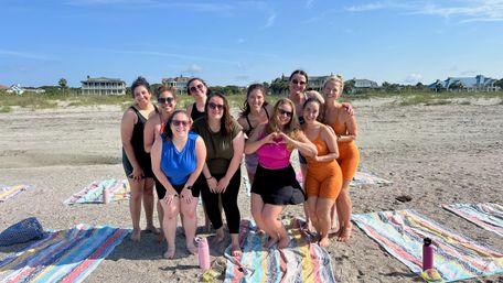 Group of smiling women in colorful activewear posing on a sandy beach over striped towels with water bottles, one making a heart with her hands, coastal homes and clear blue sky in the background.