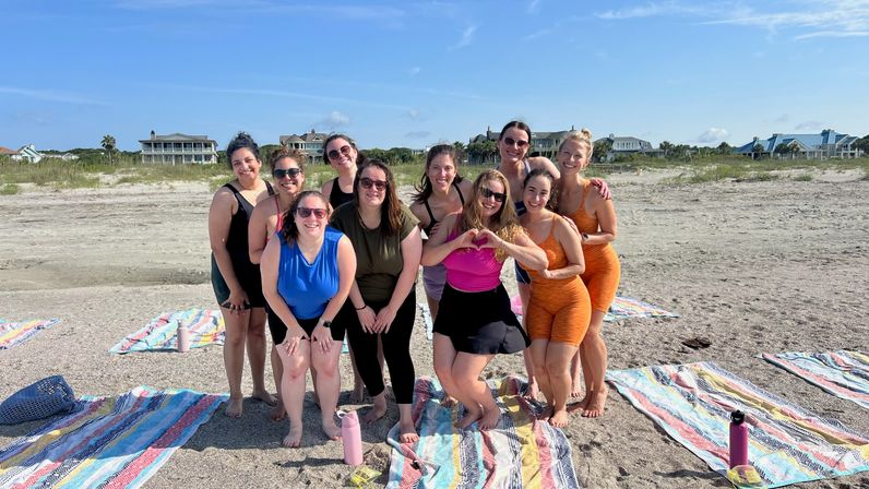 Group of smiling women in colorful activewear posing on a sandy beach over striped towels with water bottles, one making a heart with her hands, coastal homes and clear blue sky in the background.