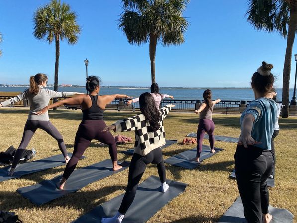 Outdoor yoga class practicing Warrior II on mats in a sunny bayfront park with palm trees, grassy lawn and calm water in the background