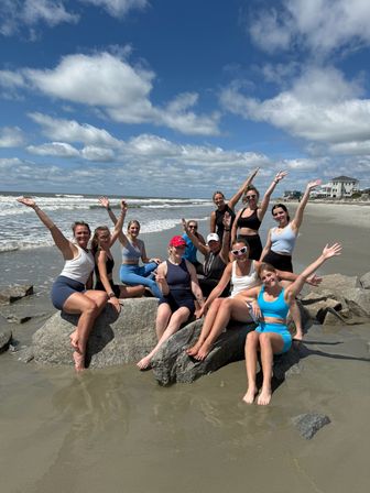 Group of friends in active swimwear posing on rocks at a sandy ocean beach under a blue sky with puffy clouds, waves rolling in and coastal houses in the background — sunny summer beach day.