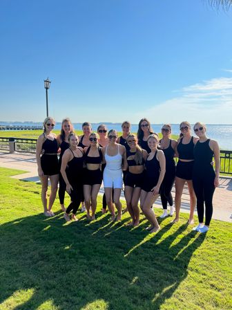 Smiling group of women in black and white workout outfits posing barefoot on a grassy waterfront promenade by a bay and pier under a clear blue sky, sunny outdoor fitness vibe.