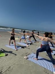 Group beach yoga class practicing Warrior II on striped mats along a sunny sandy oceanfront, waves rolling in and a pier on the horizon.