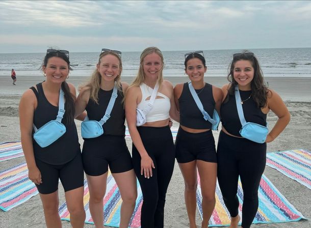 Five smiling women in black activewear wearing matching light-blue crossbody bags standing on a sandy beach by the ocean with colorful striped beach mats and a cloudy sky overhead