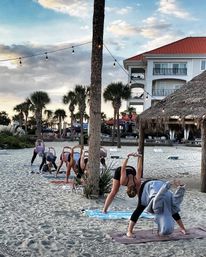Sunset beach yoga class on sandy shore with palm trees, string lights and a thatched tiki hut near a coastal resort — practitioners bent forward on mats in a relaxed, tropical scene.