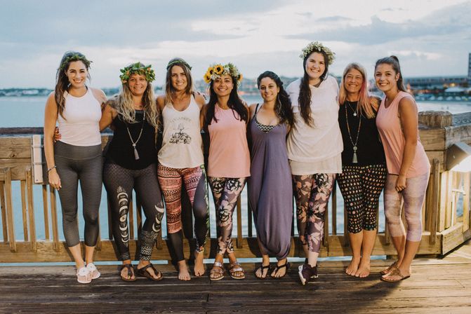 Eight women smiling on a wooden waterfront pier at sunset, wearing yoga leggings and casual tops, some with flower crowns — group photo at a seaside yoga retreat.