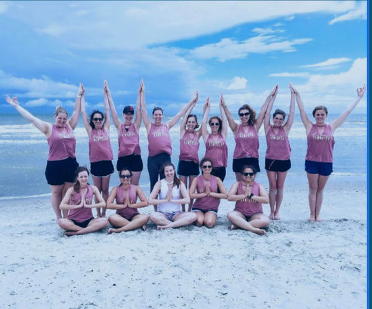 Group in matching pink tank tops striking yoga-style poses on a sandy beach, seated cross-legged and standing with arms raised against an ocean and blue sky backdrop.