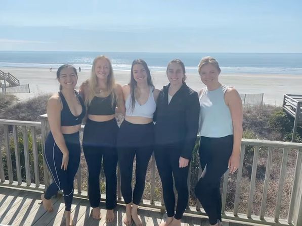Five friends in activewear standing barefoot on a wooden deck overlooking a sunny sandy beach and calm ocean, smiling for a coastal group photo.