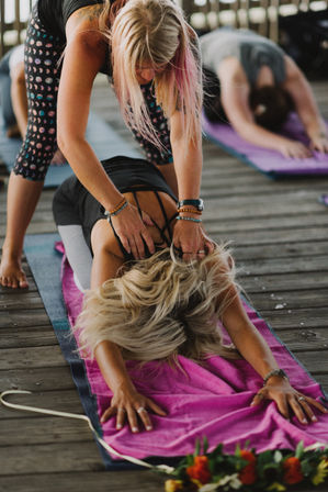 Outdoor yoga class on a wooden deck with an instructor adjusting a student’s shoulders in child’s pose on a pink towel-over-mat, other participants stretching in the background.