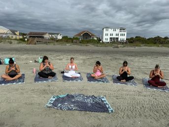 Six people seated on patterned mats practicing beach yoga meditation on a sandy shoreline with oceanfront houses and an overcast sky in the background.