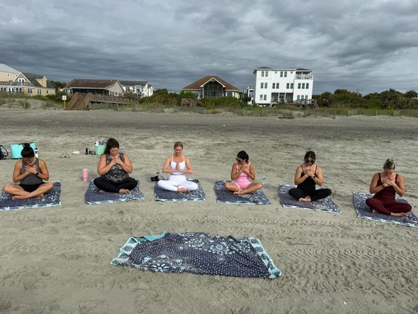 Six people seated on patterned mats practicing beach yoga meditation on a sandy shoreline with oceanfront houses and an overcast sky in the background.
