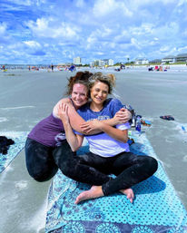 Two friends smiling and hugging on a patterned beach towel on a sandy ocean beach, with a distant pier, coastal houses and a bright cloudy sky