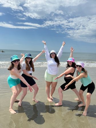 Five friends in colorful bucket hats and summer activewear posing joyfully with arms raised on a sunny sandy beach by calm ocean waves under a blue sky.