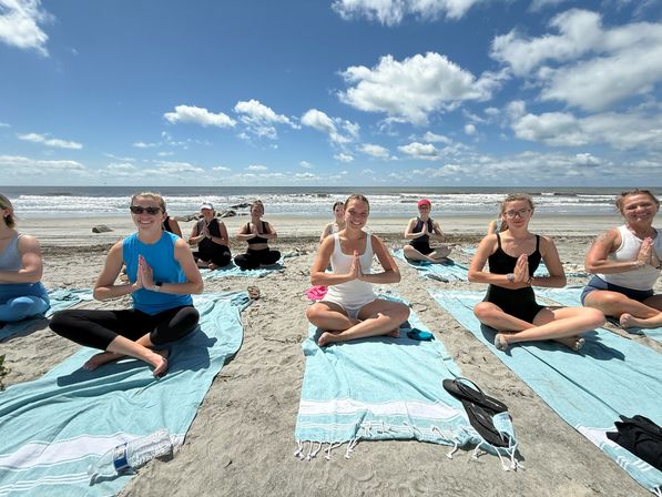Smiling group practicing beach yoga on turquoise towels, seated cross-legged in prayer pose facing the ocean with waves and a bright blue sky dotted with clouds.