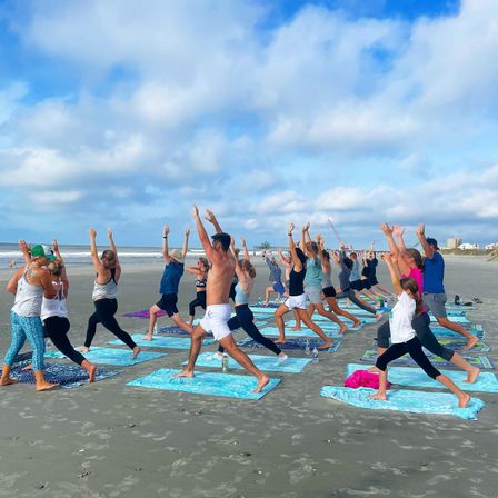 Energetic group beach yoga class on a sandy oceanfront shoreline, participants on colorful mats practicing Warrior pose with arms raised under a blue sky with scattered clouds.