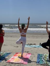Group beach yoga: person in white leggings and green cap holding tree pose on a colorful towel during a sunny seaside class with waves rolling onto the sandy shore.