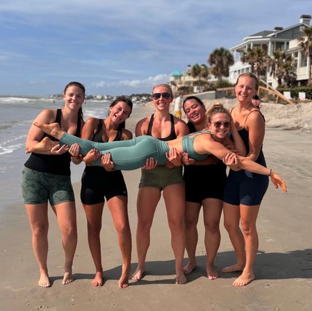 Six women in activewear laughing on a sunny beach, holding a friend horizontally near the shoreline with ocean waves and beachfront houses in the background