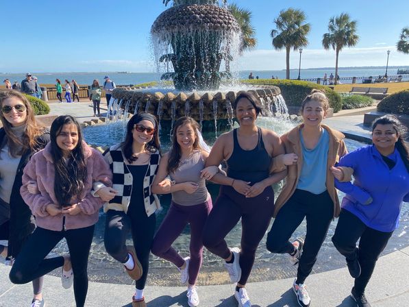 Seven friends linking arms and balancing on one leg in front of a large pineapple-style fountain on a sunny coastal promenade with palm trees and ocean views.