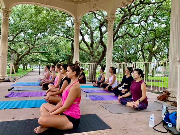Outdoor yoga class in a park pavilion under sprawling oak trees, students seated on colorful mats in meditative pose.