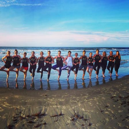 Group doing tree pose in a line on a sandy beach at sunset with ocean waves and a colorful sky in the background.