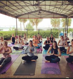 Group yoga class sitting cross-legged in namaste on colorful mats under a covered waterfront pavilion with palm trees and string lights.