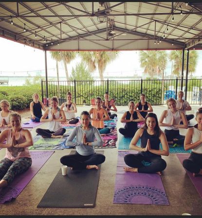 Group yoga class sitting cross-legged in namaste on colorful mats under a covered waterfront pavilion with palm trees and string lights.