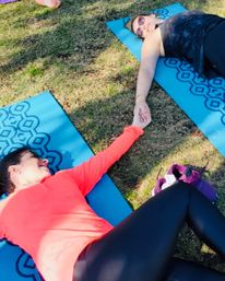 Two friends on blue yoga mats in a sunny park, lying on the grass and holding hands during an outdoor yoga cooldown.