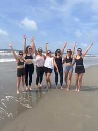 Seven women in activewear, arms raised and smiling on a sunny sandy beach by the ocean waves — barefoot summer group photo
