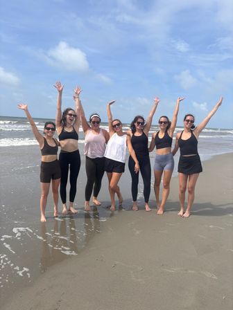 Seven women in activewear, arms raised and smiling on a sunny sandy beach by the ocean waves — barefoot summer group photo