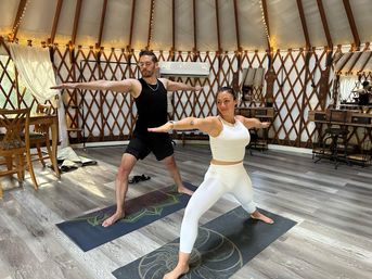 Two people practicing Warrior II on yoga mats inside a bright yurt yoga studio with wooden lattice walls, string lights, large windows and gray wood floors.