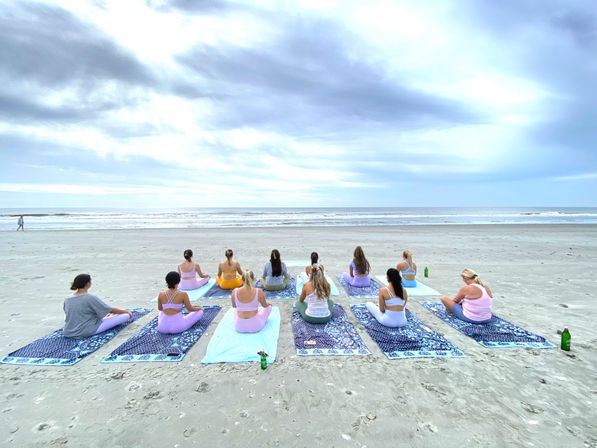 Group beach yoga class on patterned mats, people in pastel activewear seated facing the ocean under a wide cloudy sky on a sandy shoreline