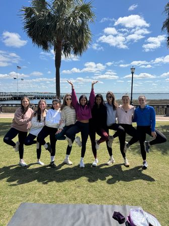 Nine friends balancing on one leg in a row under a palm tree at a sunny waterfront park, with a boardwalk, American flag and blue sky with fluffy clouds.