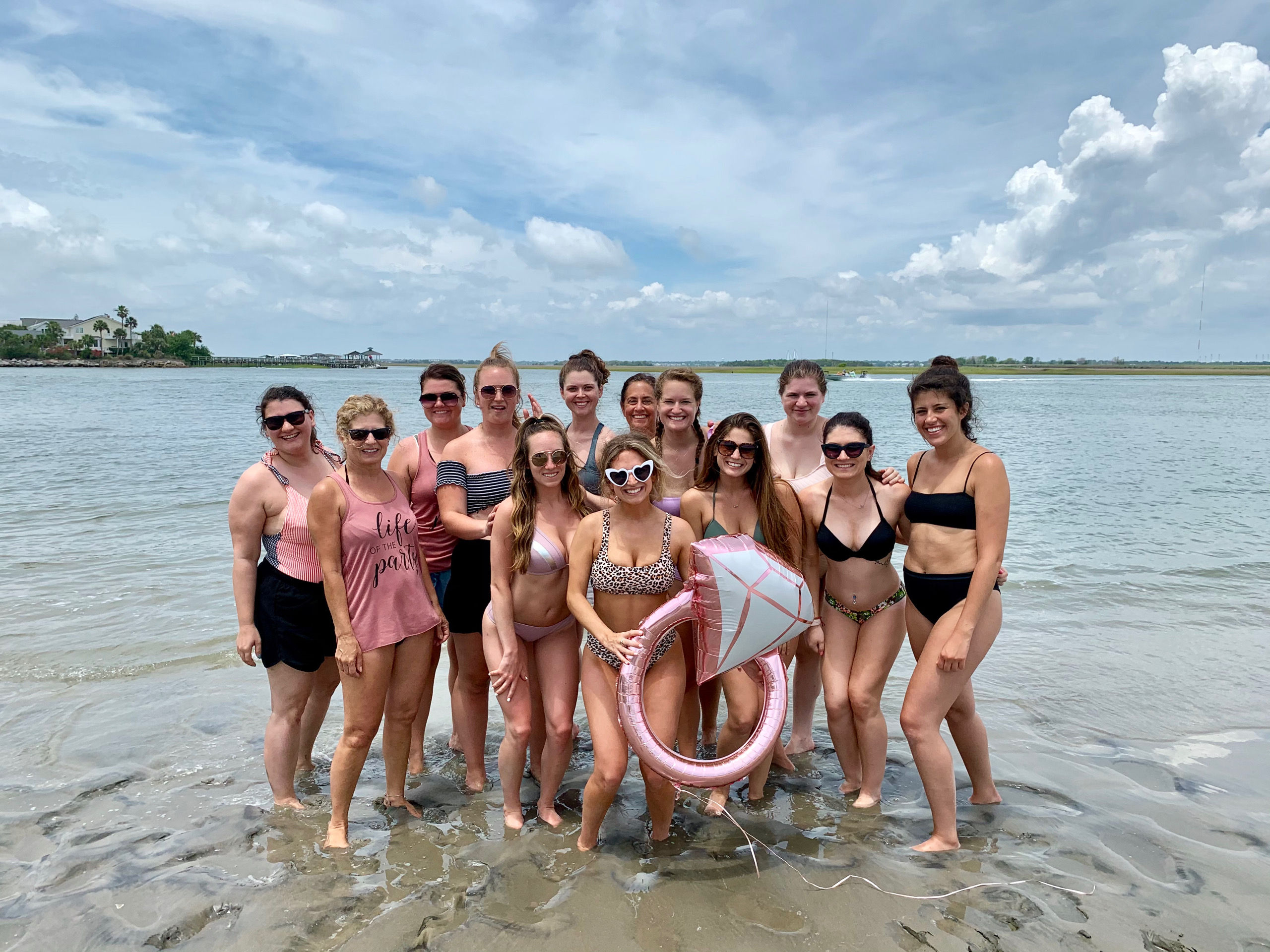 Group of women in swimsuits posing in shallow ocean water at a coastal beach on a partly cloudy day, holding a pink diamond-shaped inflatable ring for a fun beach celebration.