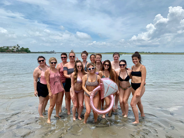 Group of women in swimsuits posing in shallow ocean water at a coastal beach on a partly cloudy day, holding a pink diamond-shaped inflatable ring for a fun beach celebration.