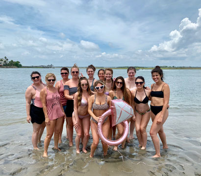 Group of women in swimsuits posing in shallow ocean water at a coastal beach on a partly cloudy day, holding a pink diamond-shaped inflatable ring for a fun beach celebration.