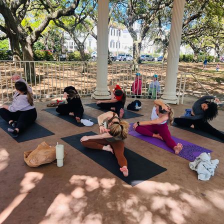 Group yoga class on mats in a park gazebo under sprawling oak trees, participants stretching in seated side-bend poses — sunny, shaded outdoor workout.