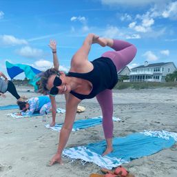 Smiling woman in sunglasses doing a standing bow yoga pose on a towel at a sandy beach yoga class, wearing mauve leggings and a black tank, with other participants and beachfront houses under a blue sky.