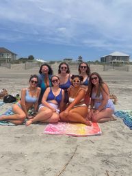 Seven friends in swimsuits and sunglasses sitting on colorful towels on a sunny sandy beach with dunes and oceanfront houses in the background — cheerful summer beach day.