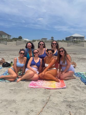 Seven friends in swimsuits and sunglasses sitting on colorful towels on a sunny sandy beach with dunes and oceanfront houses in the background — cheerful summer beach day.