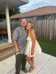 Two people smiling on a residential backyard patio at dusk — one wearing a chef jacket and bandana, the other barefoot in a short cream satin dress, with a wooden fence, deck, and lawn in the background.