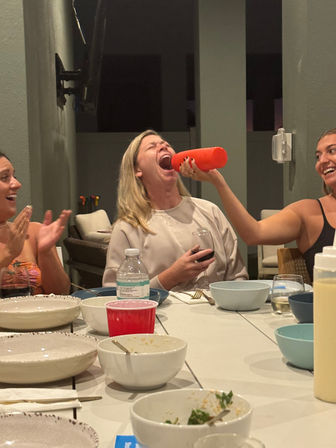 Three friends at a casual patio dinner party, laughing as one woman playfully pours from a red bottle into another’s open mouth, table scattered with bowls, plates, wine glasses and a red solo cup.