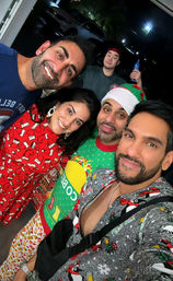 Holiday pajama-party selfie of five friends in Christmas sweaters, Santa hat and festive pajamas, smiling at a home's open front doorway at night.