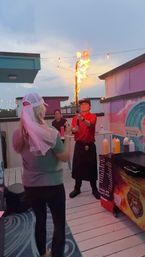 Rooftop patio at dusk with a server holding a flaming torch over a food cart while a guest wearing a white veil watches under string lights.