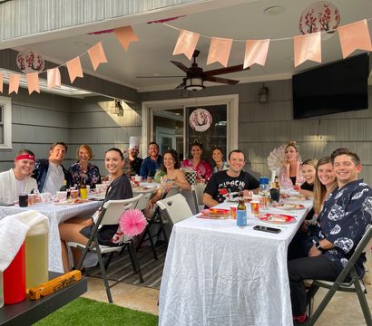 Group of friends at a covered backyard patio party — outdoor dining on long white-tablecloth tables with pink bunting, paper lanterns, drinks and folding chairs, smiling and enjoying the gathering.