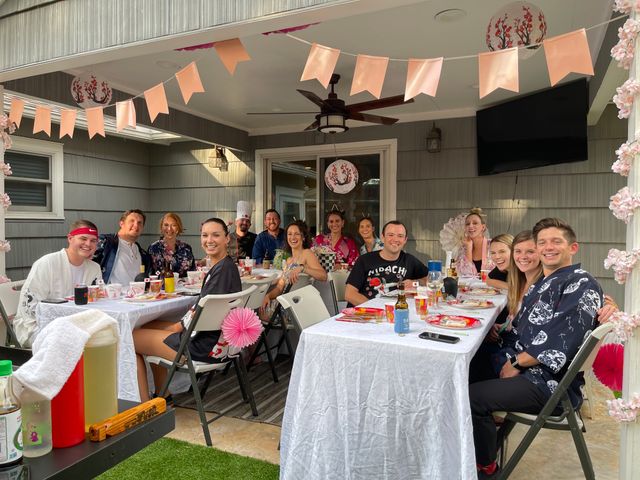 Group of friends at a covered backyard patio party — outdoor dining on long white-tablecloth tables with pink bunting, paper lanterns, drinks and folding chairs, smiling and enjoying the gathering.