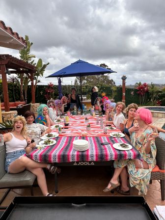 Group of friends wearing colorful wigs sharing a poolside brunch at a long striped table on a backyard patio under a blue umbrella and cloudy sky