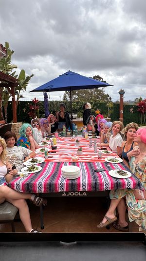 Group of friends wearing colorful wigs sharing a poolside brunch at a long striped table on a backyard patio under a blue umbrella and cloudy sky