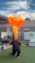 Backyard fire breather kneels on artificial turf, holding torches and exhaling a towering orange flame plume in a residential patio with a propane tank and tiled roof under a blue sky.