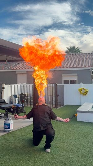 Backyard fire breather kneels on artificial turf, holding torches and exhaling a towering orange flame plume in a residential patio with a propane tank and tiled roof under a blue sky.