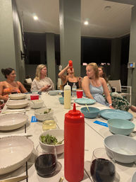 Casual outdoor patio dinner at night with a group of women around a long table of bowls, salads, wine glasses and a prominent red squeeze bottle, one woman tipping a bottle to drink.